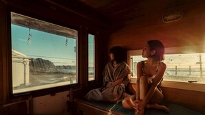 L'interno di un treno con due passeggeri seduti accanto a grandi finestre, rivestimenti in legno e vista su un ponte e un paesaggio.
