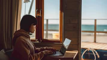 Une personne est assise à une table en bois près d’une fenêtre, devant un ordinateur portable, et la vue donne sur la plage.