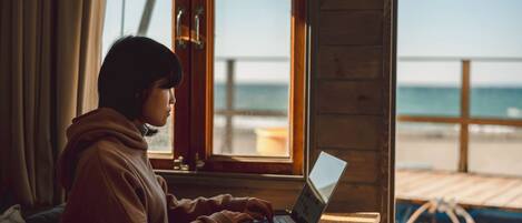 A person using a laptop at a wooden table by a window with a view of the beach.