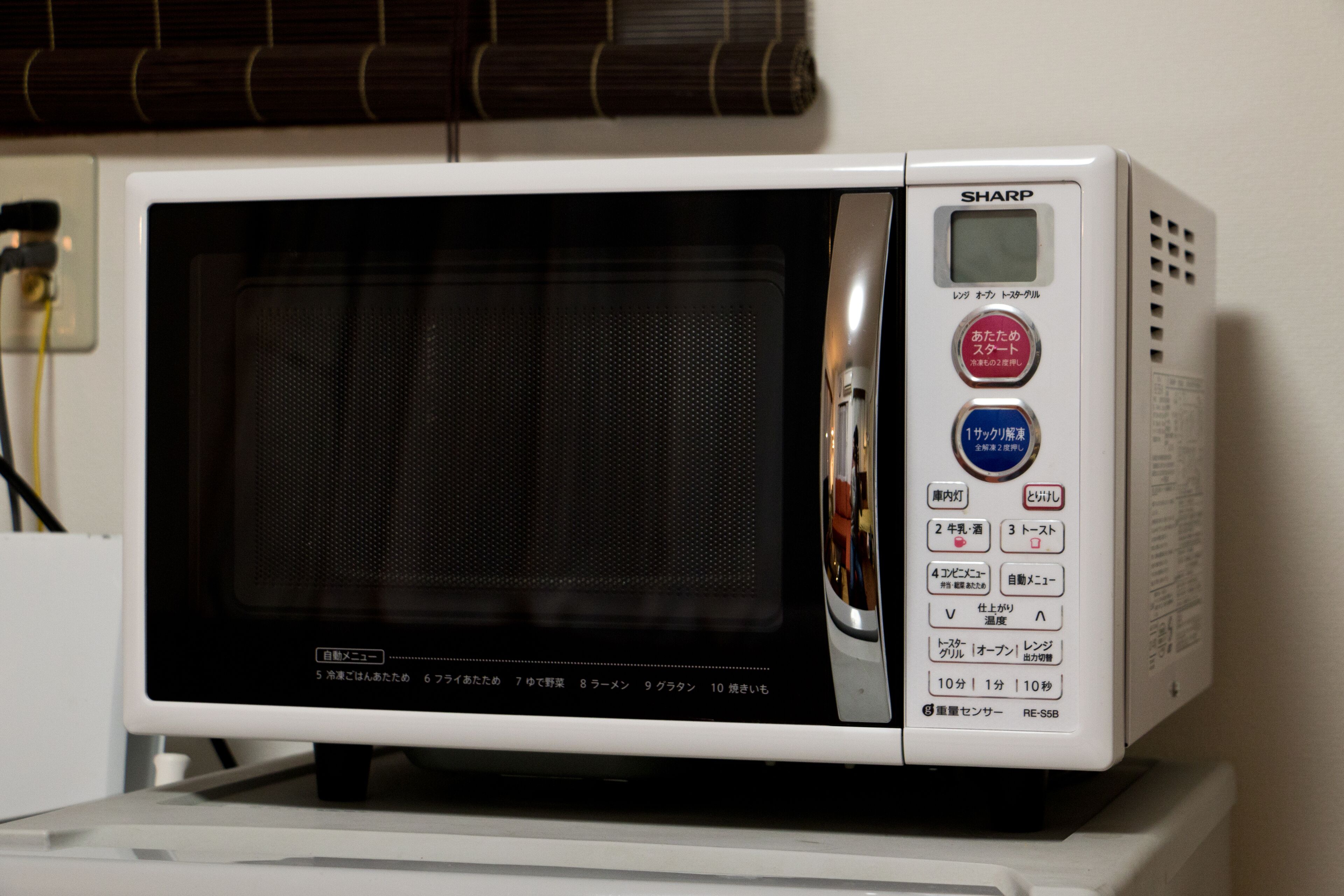 Un horno microondas Sharp blanco con pantalla digital y varios botones, incluyendo un botón de encendido rojo y un botón de inicio azul.