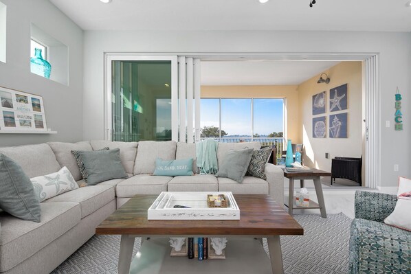 A modern living room with a beige sofa, a wooden coffee table, and a view of the ocean through large sliding glass doors. - Exclusive Anna Maria Sound Waterfront Retreat with Shared Pool & Sunset Serenity (Bradenton)