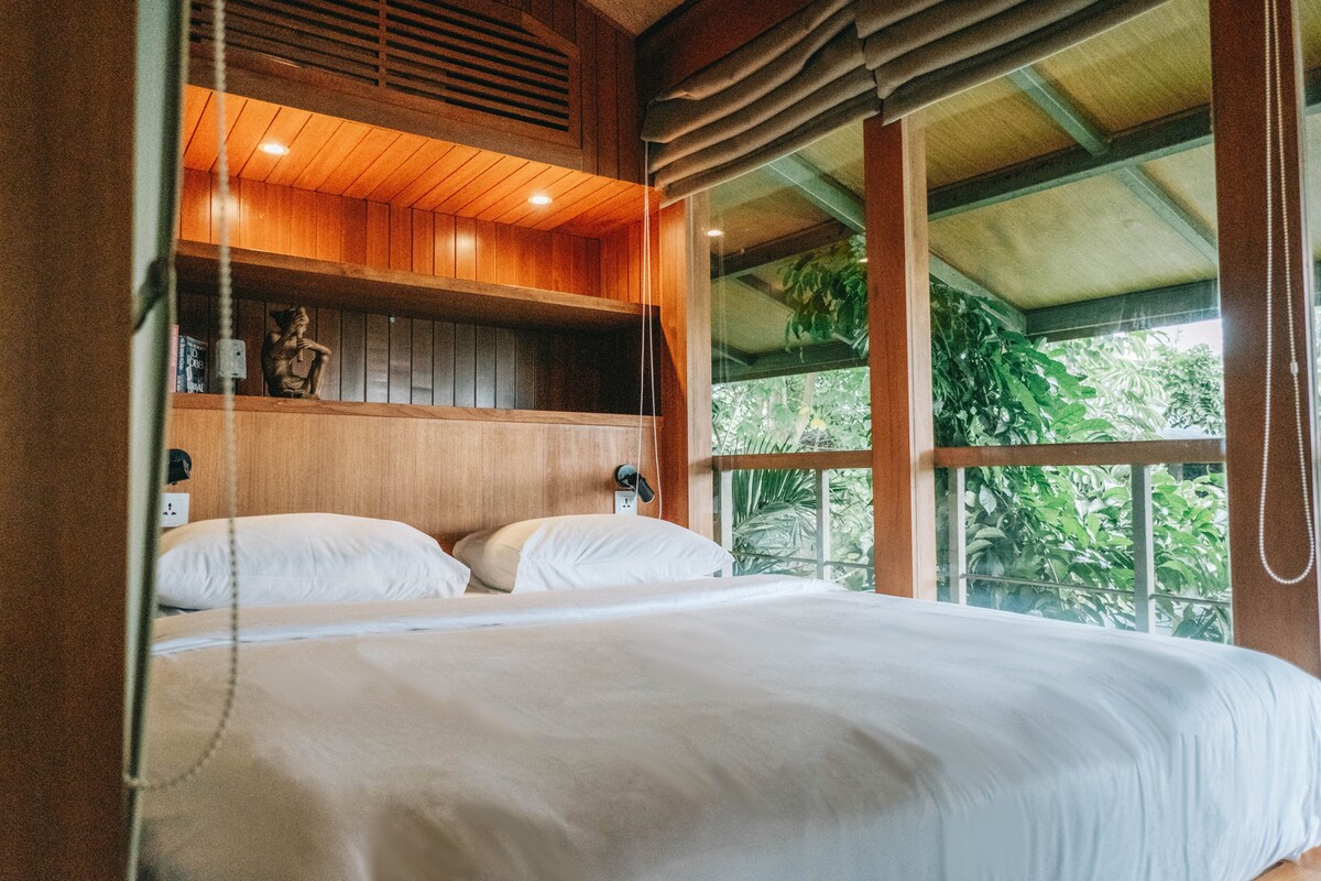 Une chambre avec un plafond en bois, un lit recouvert de draps blancs, une table de chevet et une fenêtre donnant sur un paysage verdoyant.