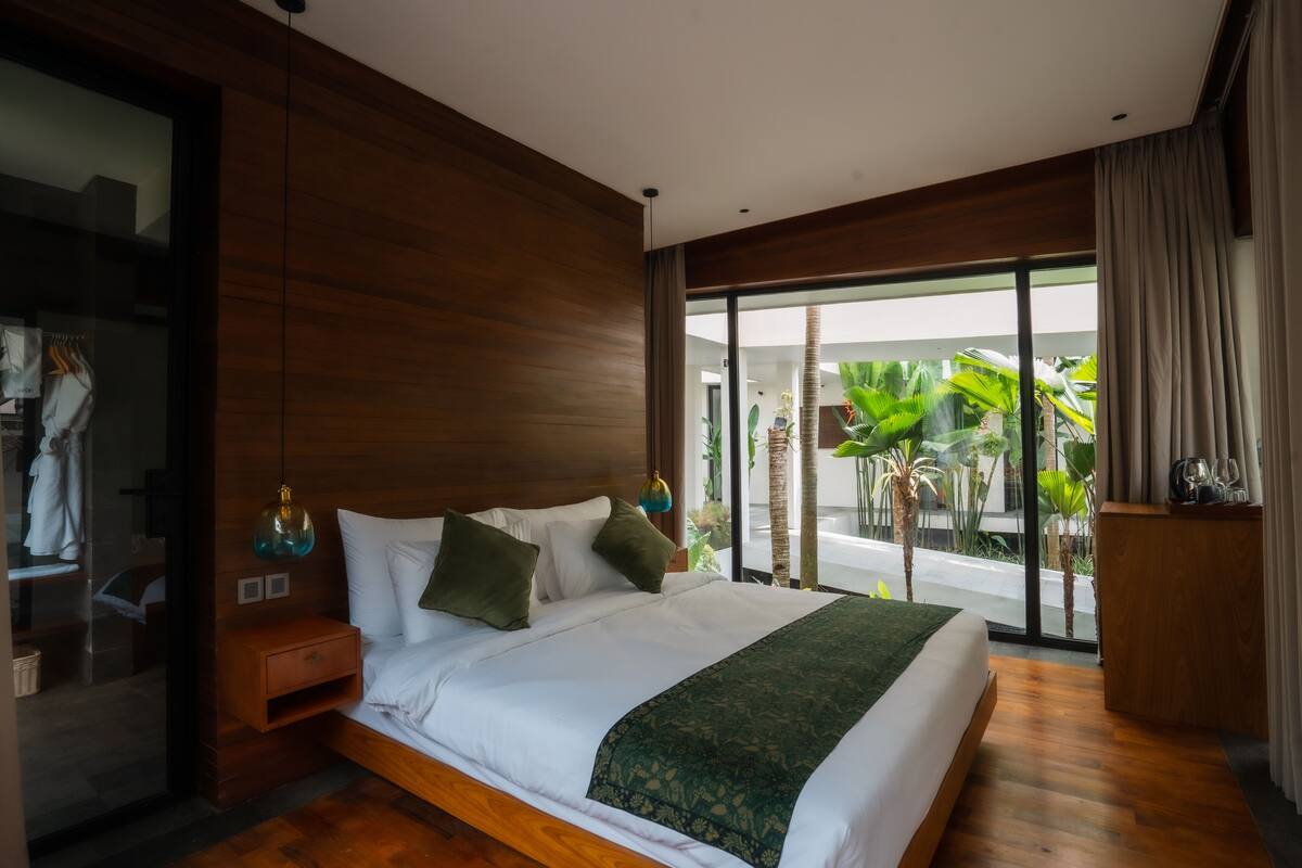 Ein modernes Hotelzimmer mit einem großen Bett, Holzboden, einer Glastür, die auf einen Balkon führt, und Blick auf tropische Pflanzen.