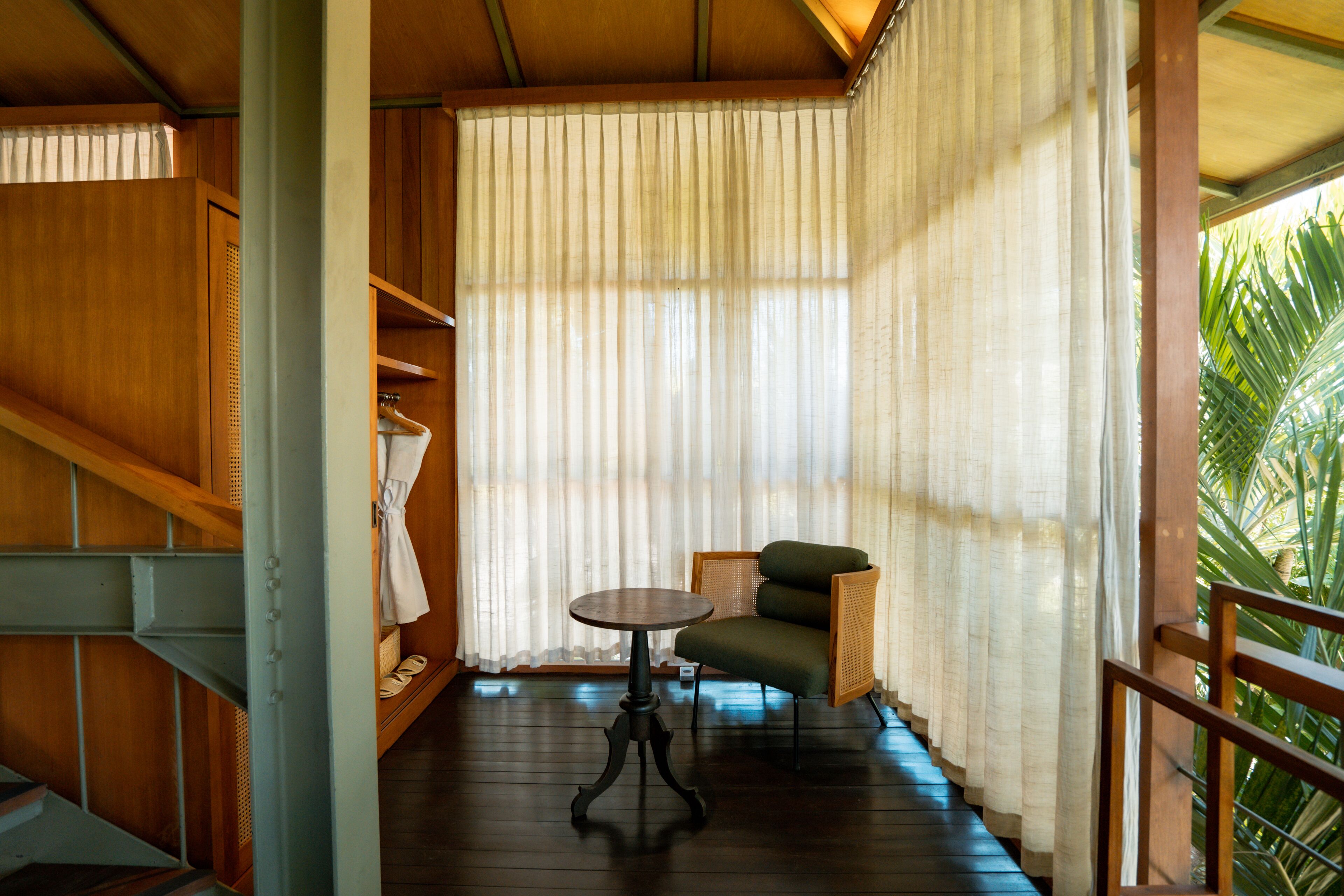 Una escalera de madera que lleva a una habitación con piso de madera, una mesa redonda, una silla verde y una ventana con cortinas traslúcidas.