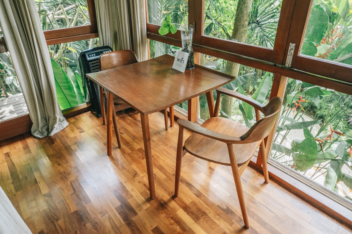 Une table et des chaises en bois sont disposées dans une pièce dotée de grandes fenêtres, d’un plancher en bois et d’un appareil de climatisation visible.