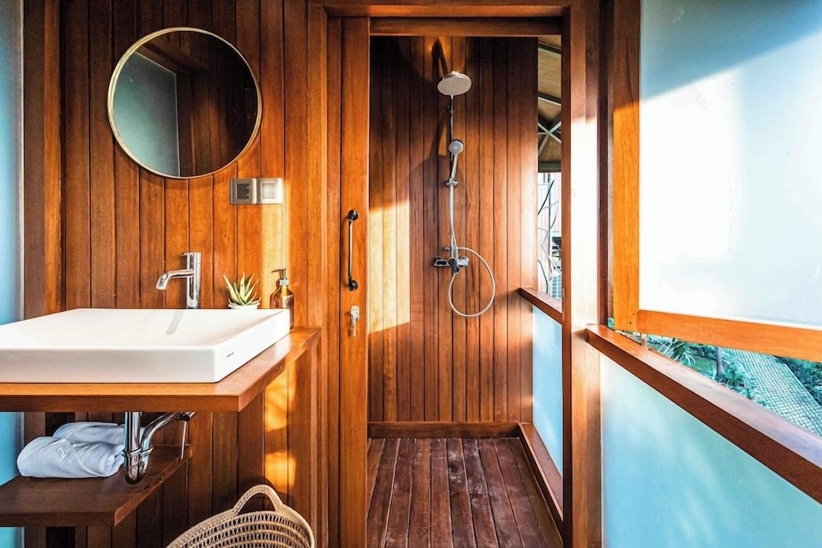 Une salle de bain en bois avec un lavabo, un miroir, une douche et une fenêtre donnant sur l’extérieur.