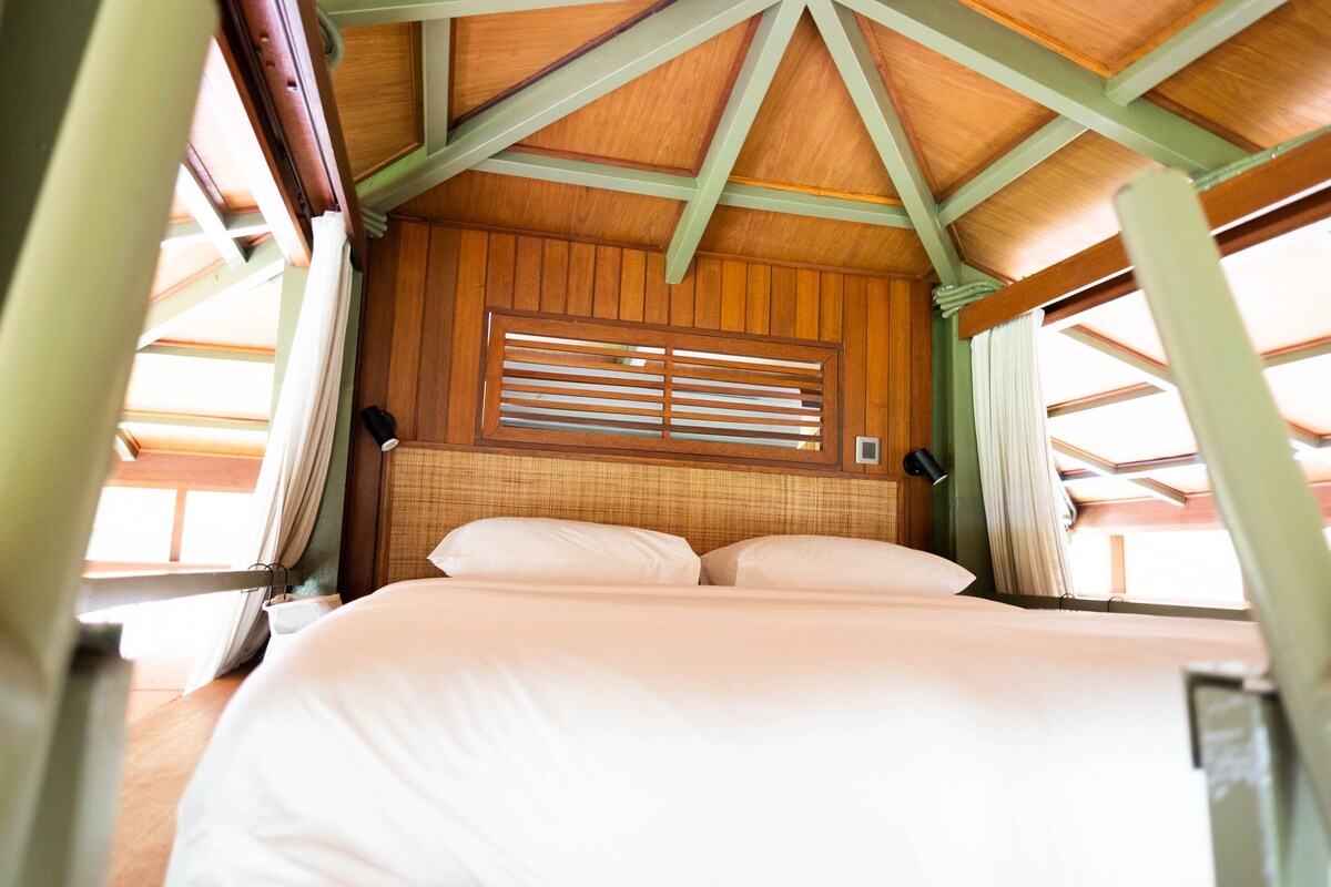 A room with a bed, wooden ceiling beams, a window with blinds, and a wall-mounted lamp.