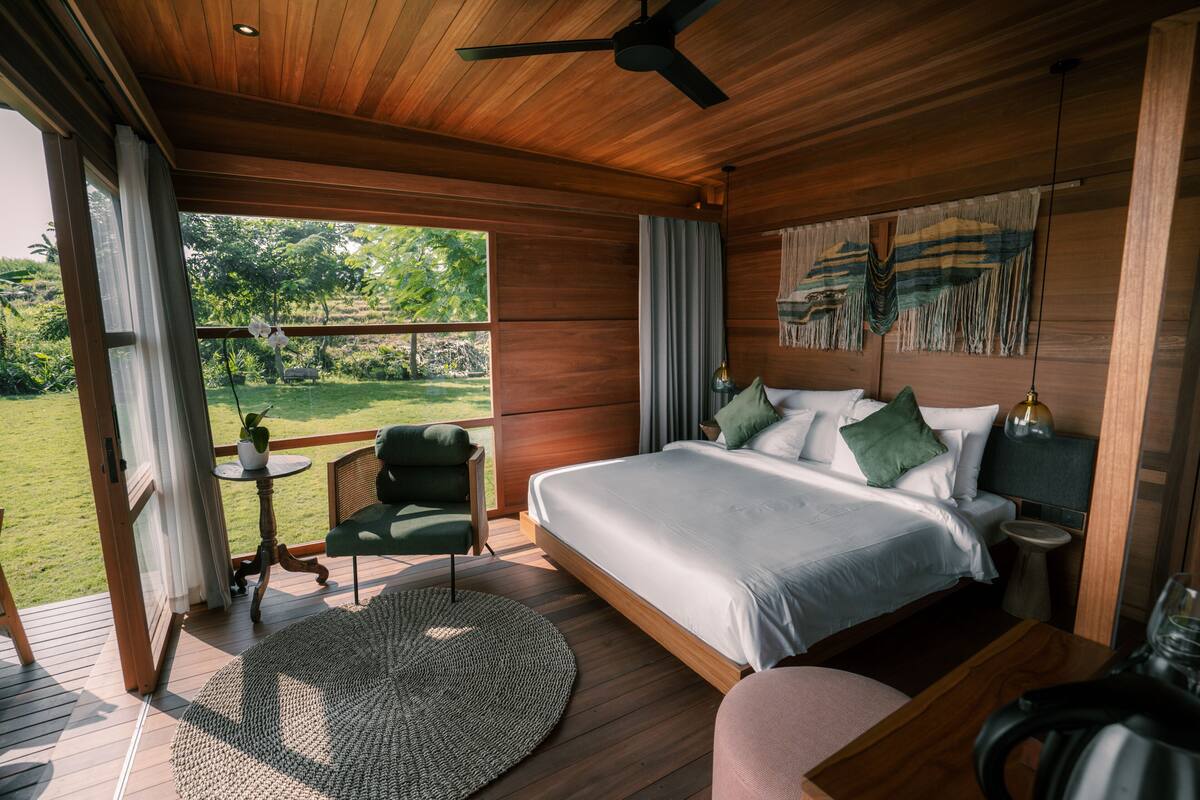 Ein Schlafzimmer mit einem großen Bett, einem Deckenventilator, einem kleinen Tisch mit einer Pflanze und Blick ins Freie.