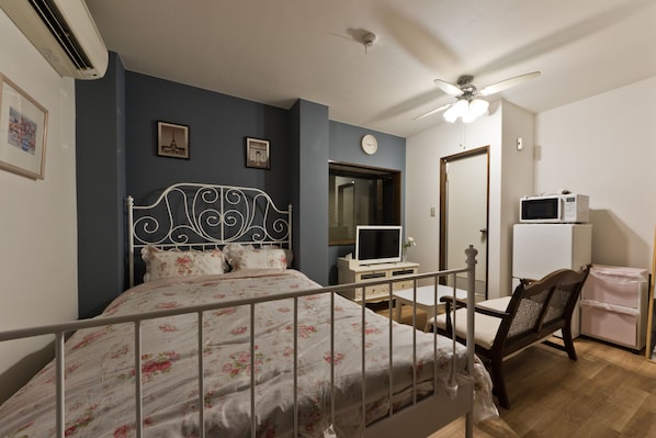 A bedroom with a bed, a desk, a chair, a ceiling fan, a clock, and a refrigerator.