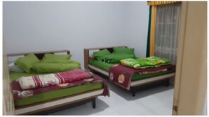 Duas camas de solteiro com roupa de cama verde e estruturas de madeira em um quarto com paredes brancas e uma janela com cortinas amarelas.