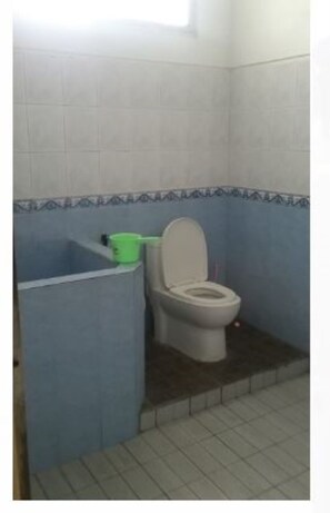 A small bathroom with a toilet, a green bucket, and a blue-tiled shower area. - Villa Darsono (Batu)