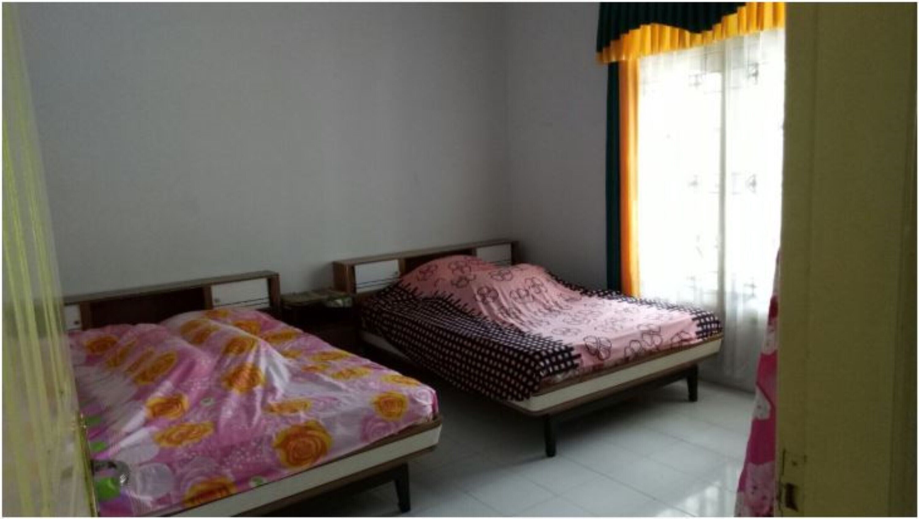 Two single beds with wooden frames and headboards, a window with yellow curtains, and a tiled floor.