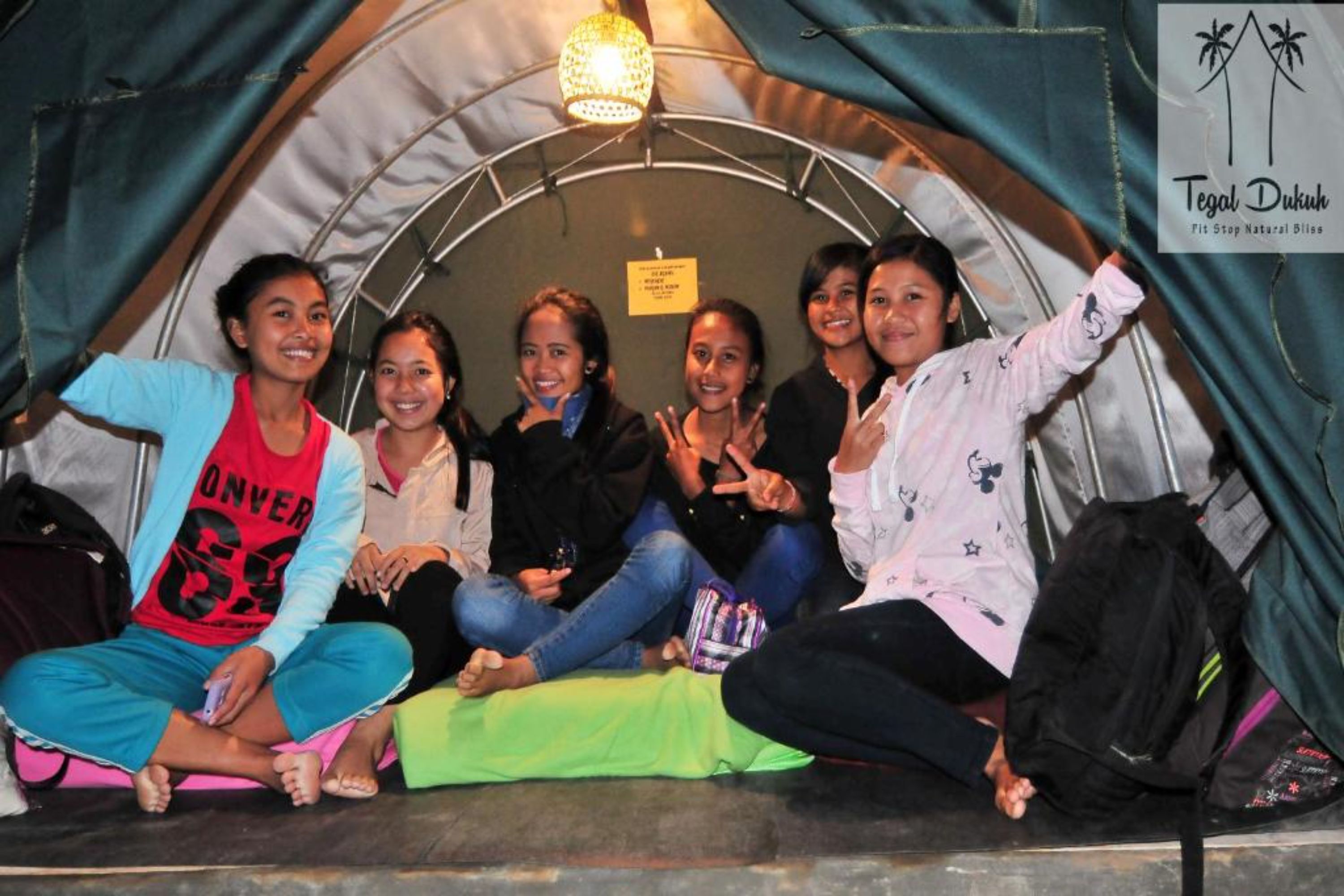 A group of people sitting inside a tent-like structure with a sign that reads "Tegal Dukuh Fit Stop Natural Bliss".