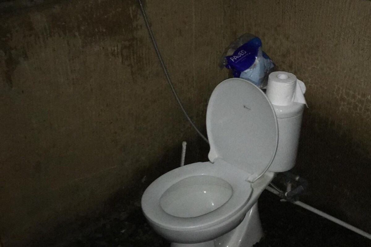 A toilet in a dimly lit, confined space with a visible pipe and a bag of tissues on the tank.