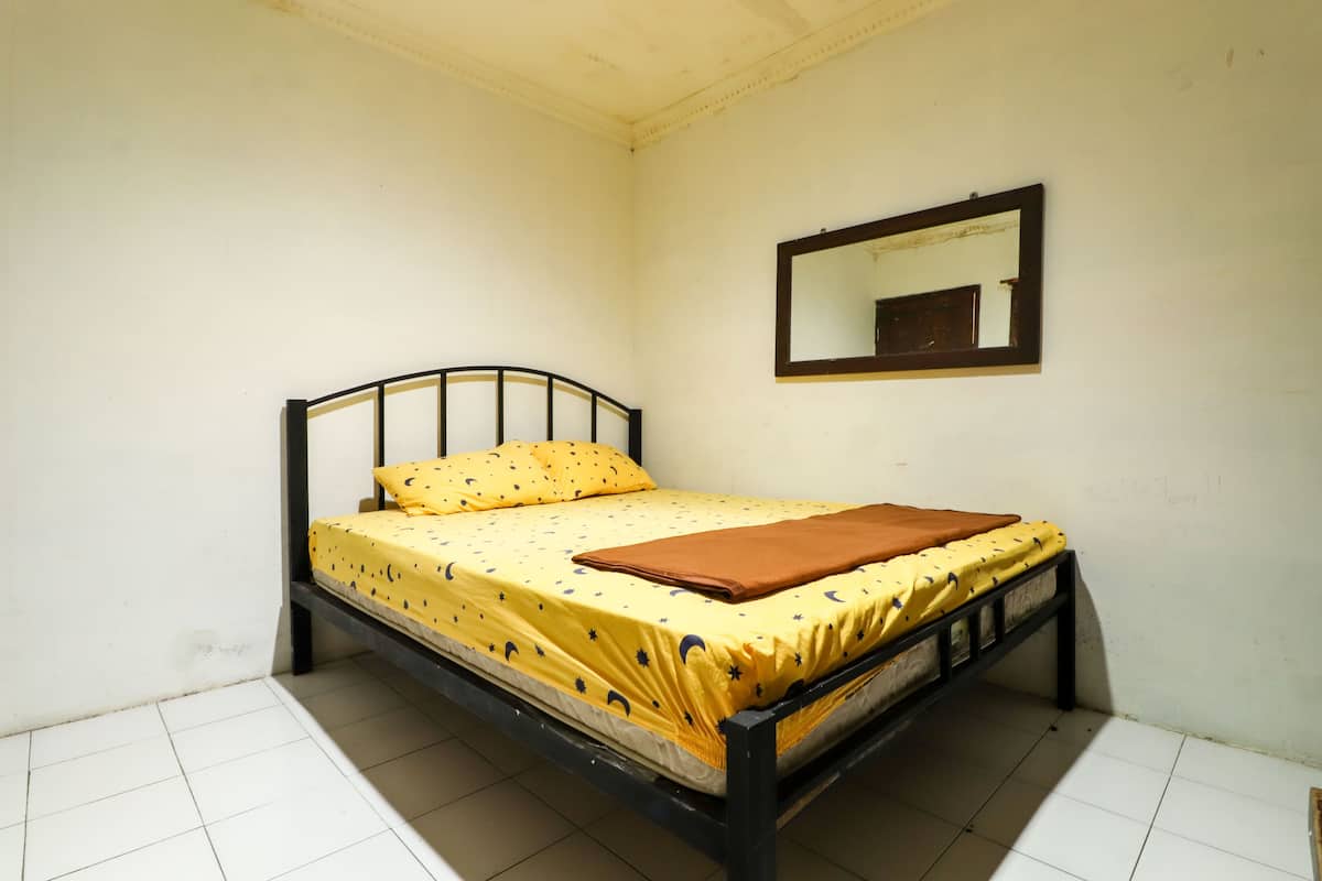 A single bed with a yellow bedspread and a brown blanket, a small mirror on the wall, and white tiled flooring.