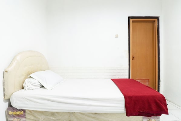 Una cama individual con colchón blanco, cabecera color beige y manta roja. Se ve una puerta de madera al fondo.