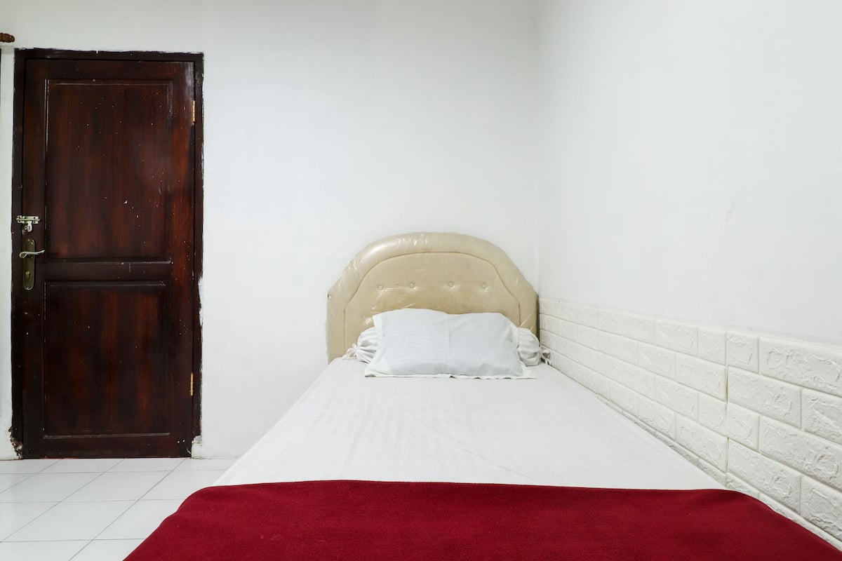 A single bed with a white mattress and a red blanket, a beige upholstered headboard, and a wooden door.