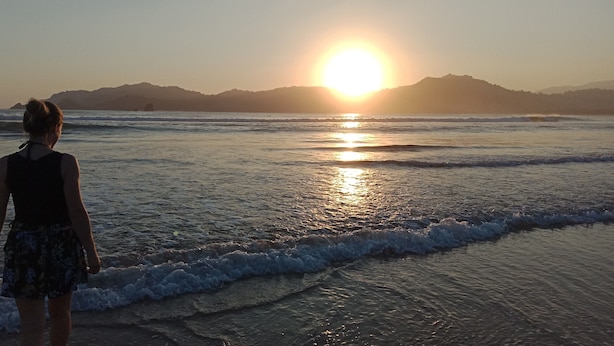A person standing on a beach facing the ocean with the sun setting in the background. - Cheap Inn - Hostel (Banyuwangi)