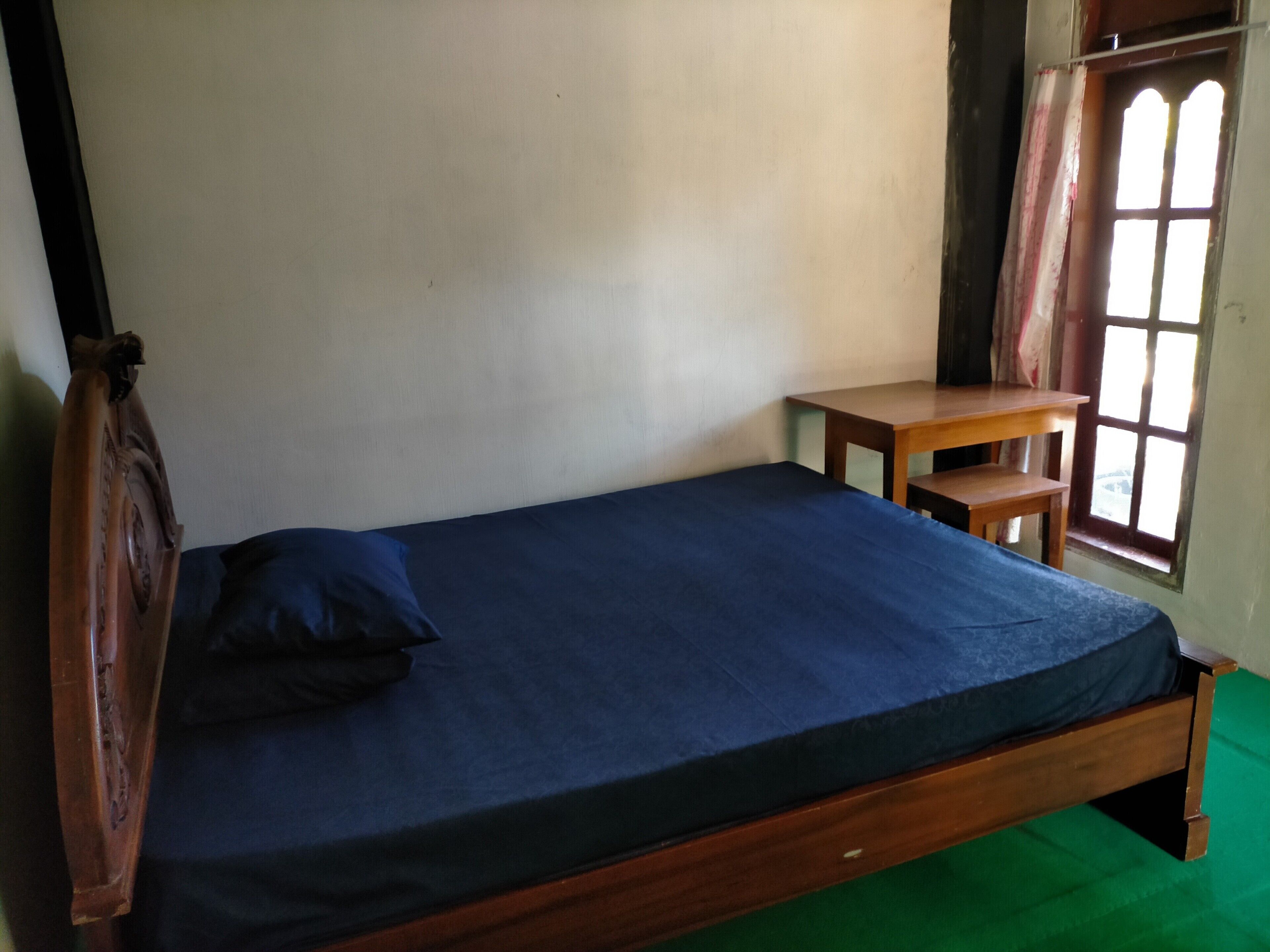 A single wooden bed with a blue mattress and a pillow, a small wooden table, and a window with a view of the outside.