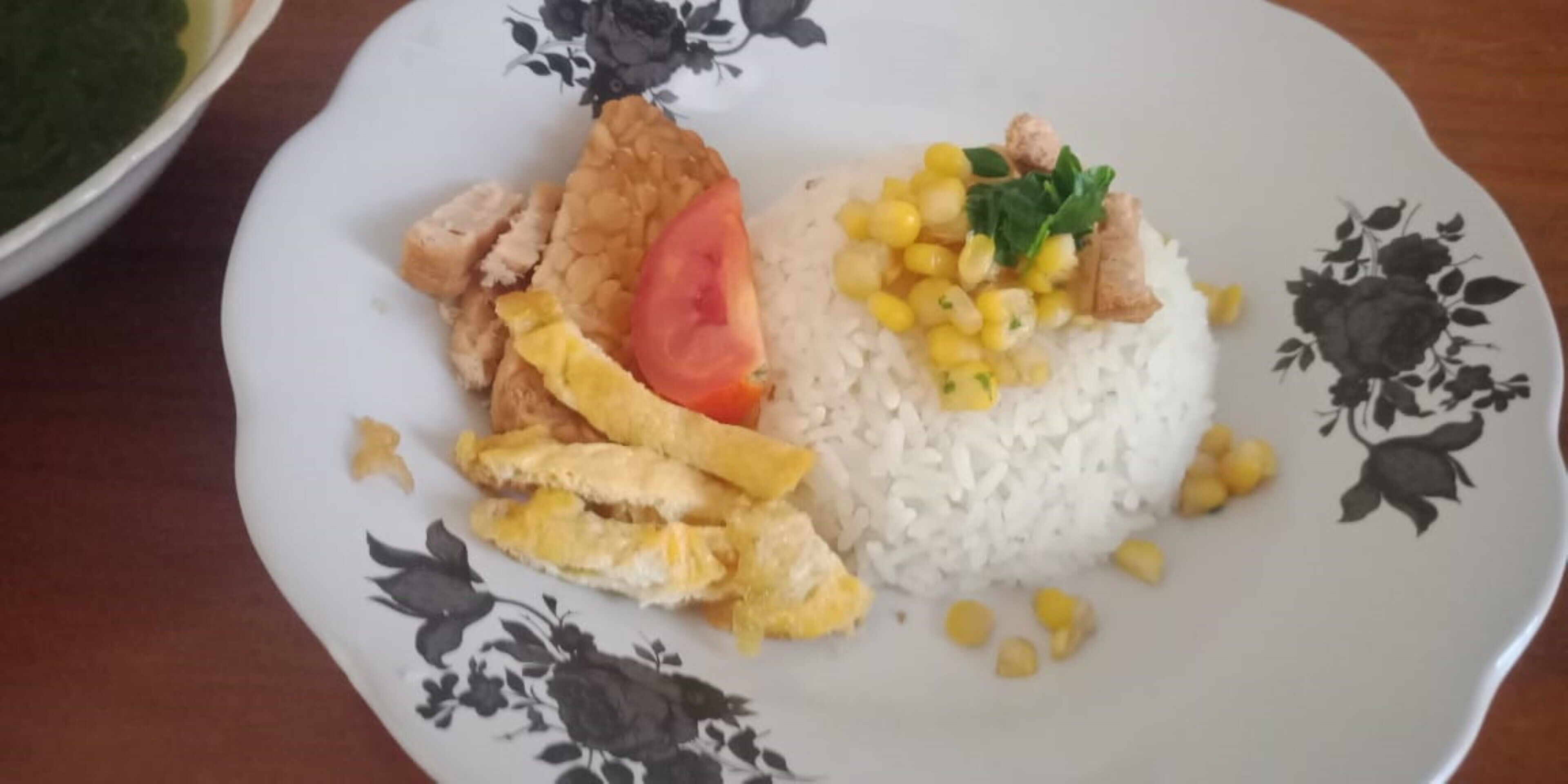 A plate with white rice, corn, a slice of tomato, scrambled eggs, and a piece of chicken.