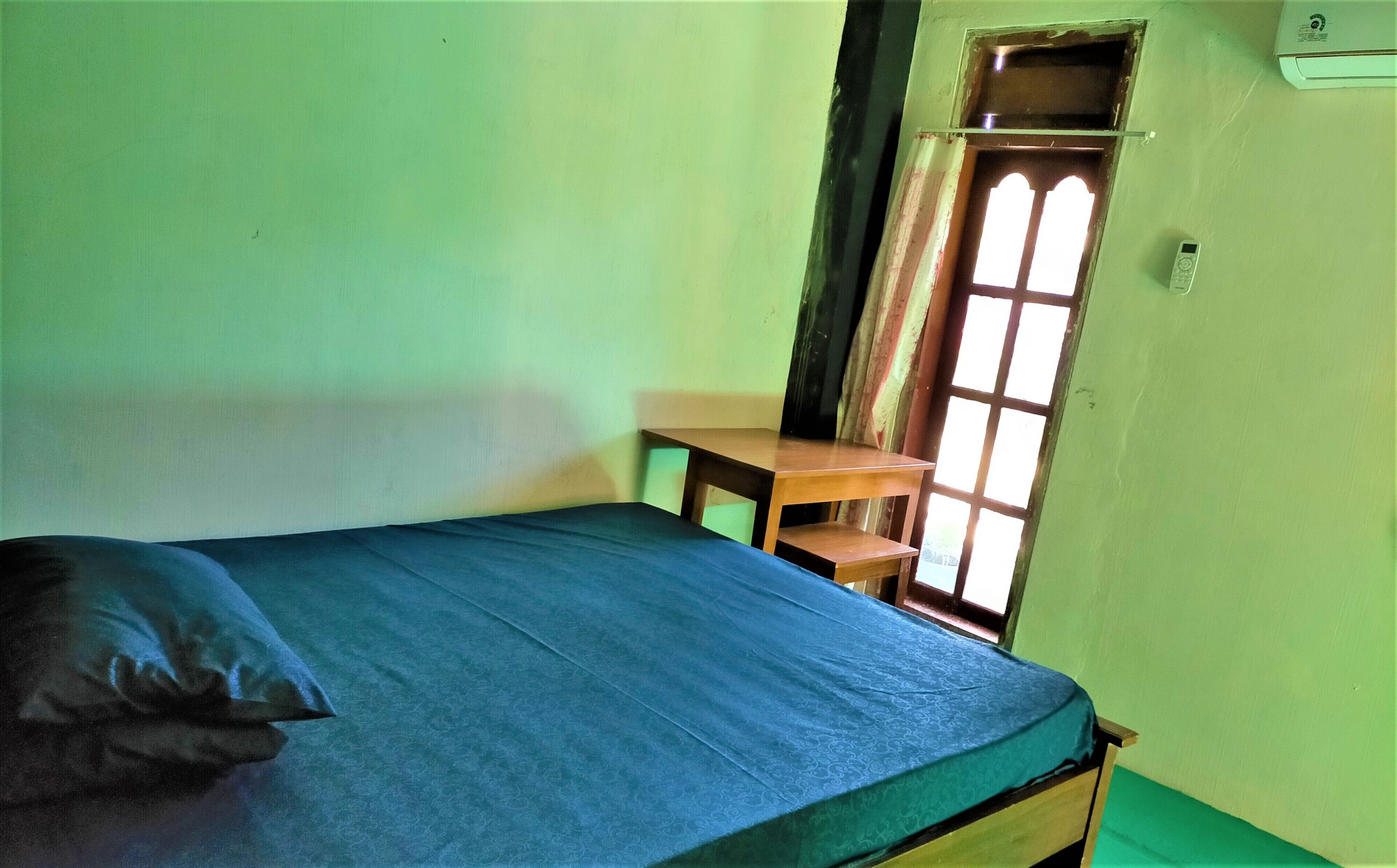 Un lit simple avec un matelas bleu et un oreiller, une table en bois et une fenêtre avec un rideau.
