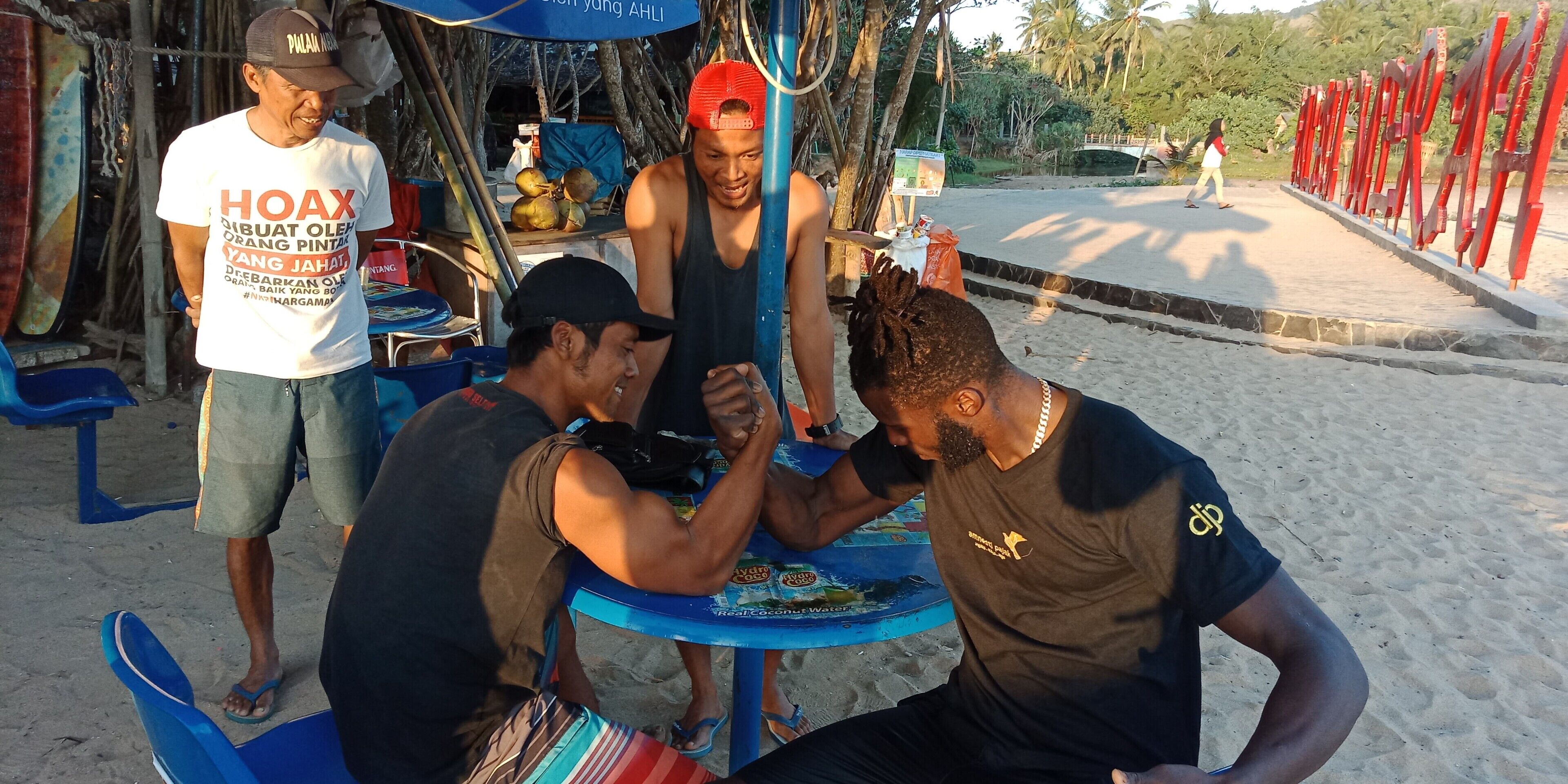 A group of people sitting around a table with a blue umbrella, a signboard, and a beach setting in the background.