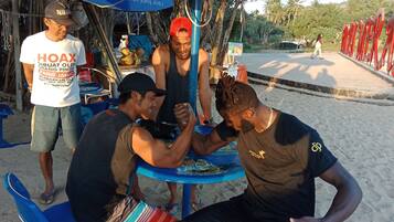 A group of people sitting around a table with a blue umbrella, a signboard, and a beach setting in the background.
