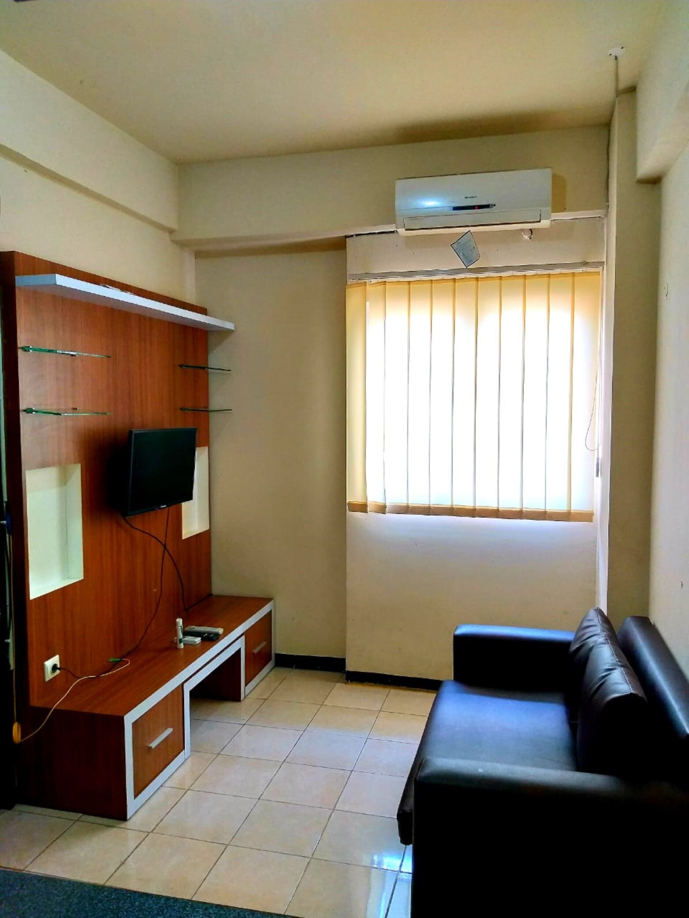 A living room with a wooden entertainment unit, a flat-screen TV, a window with blinds, a black leather sofa, and an air conditioning unit.
