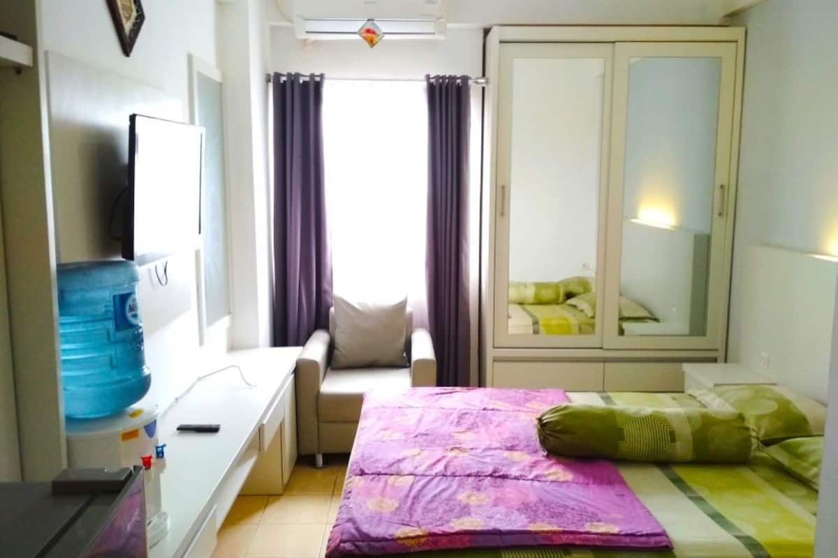 Habitación de hotel pequeña con cama, escritorio, silla, armario y un ventanal con cortinas.