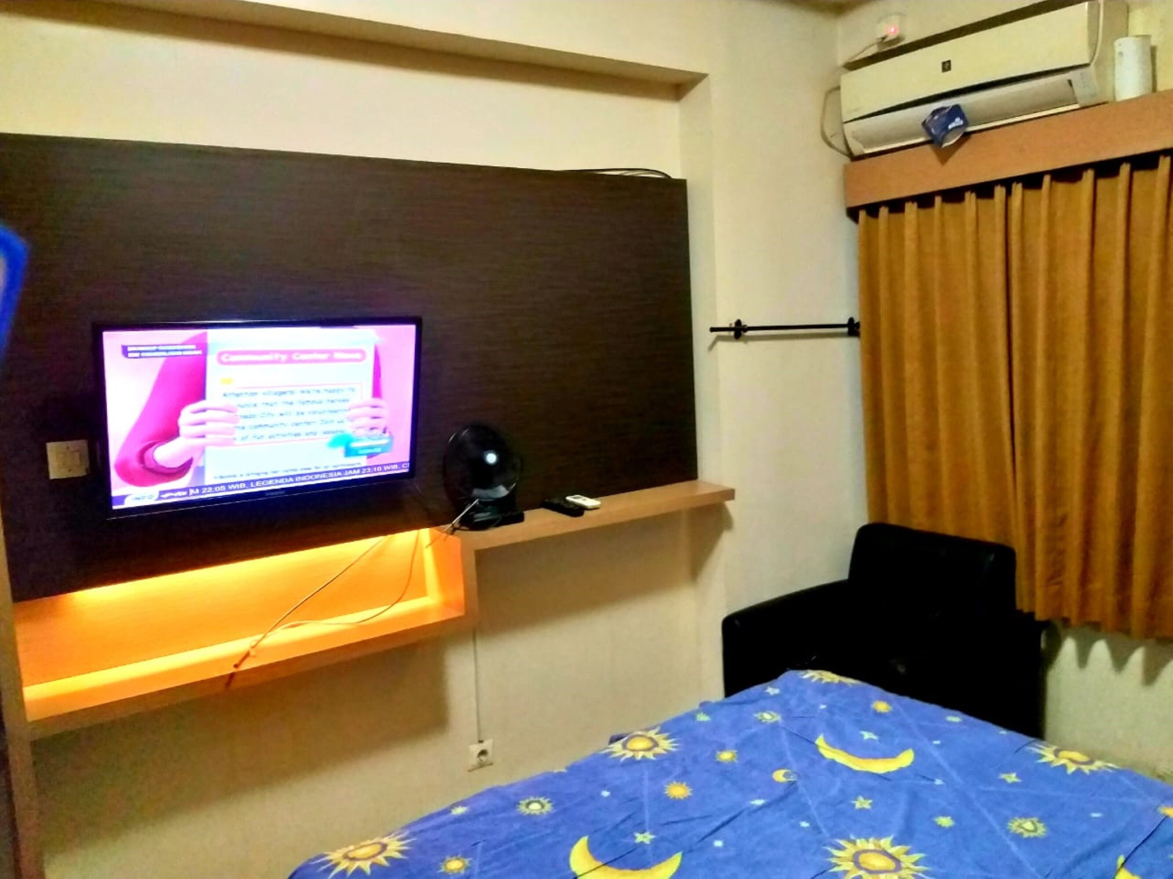 A hotel room with a television mounted on the wall, a bed with a blue bedspread featuring sun and moon patterns, a small table, and an air conditioning unit above the bed.
