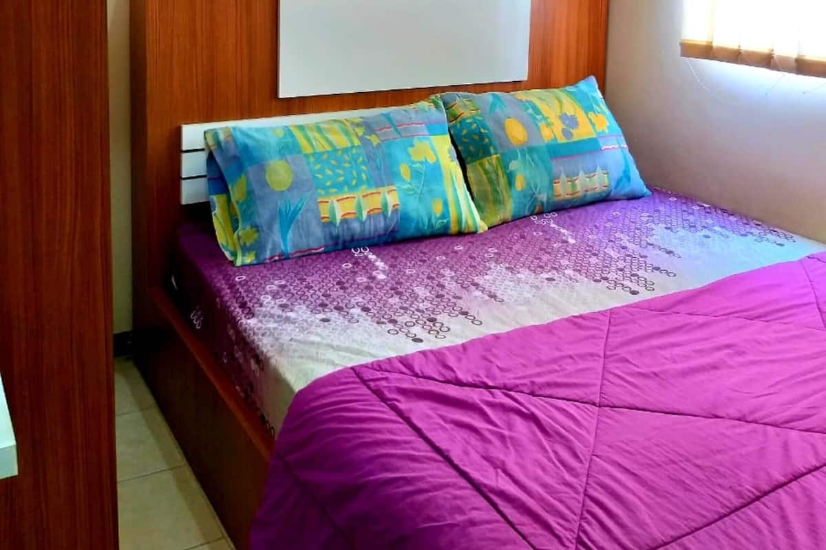 Un dormitorio con cabecera de madera, una cama con colcha morada y dos almohadas coloridas, una ventana con persianas y una puerta de madera.
