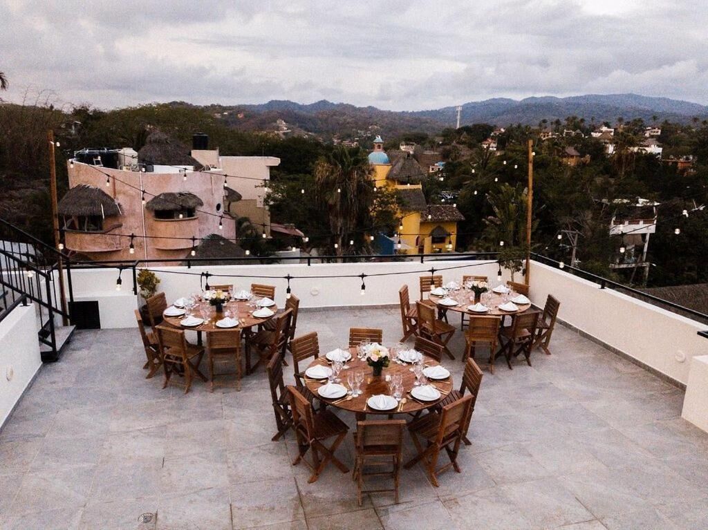 A rooftop terrace with multiple round tables set for dining, overlooking a mountainous landscape with buildings and greenery.