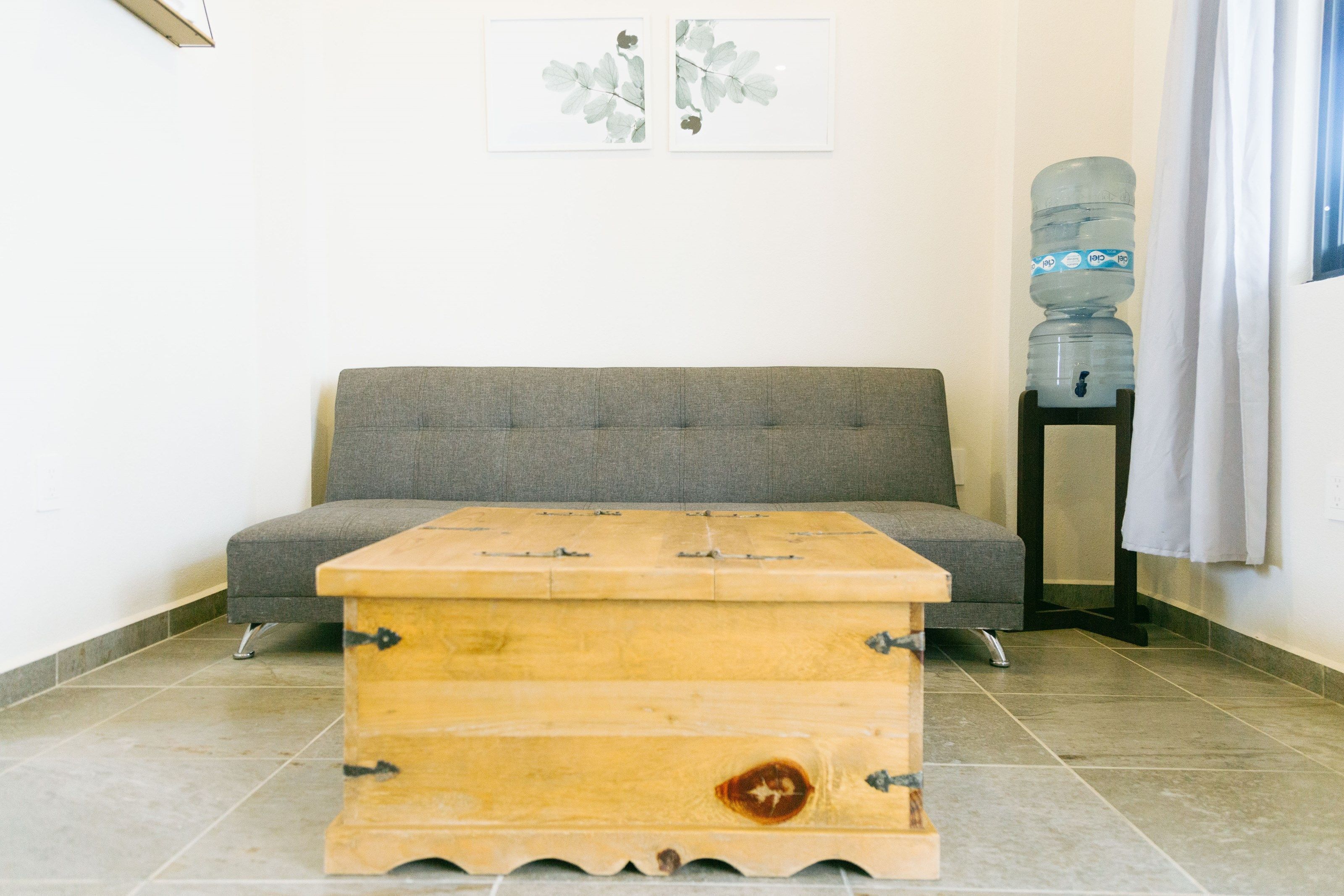A room with a grey sofa, a wooden chest, a water dispenser, and a framed artwork on the wall.