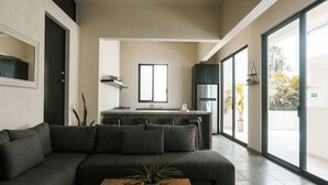 Uma sala de estar moderna com um sofá escuro, uma mesa de centro de madeira e uma área de cozinha com uma janela grande.