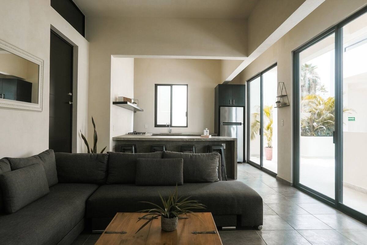 Ein modernes Wohnzimmer mit einer dunkelgrauen Couch, einem Holztisch und einer Küchenzeile mit großem Fenster.