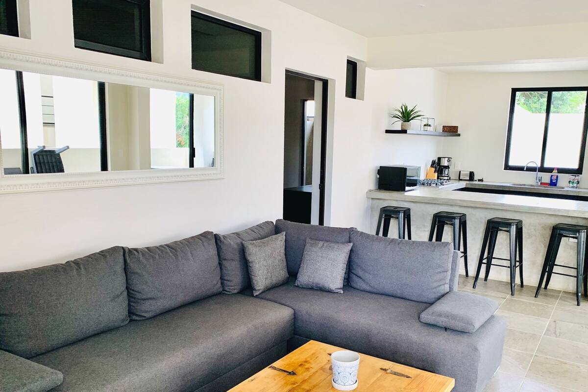 Ein modernes Wohnzimmer mit einem grauen Ecksofa, einem Holztisch und einer Küchenzeile mit Bar und Hockern.