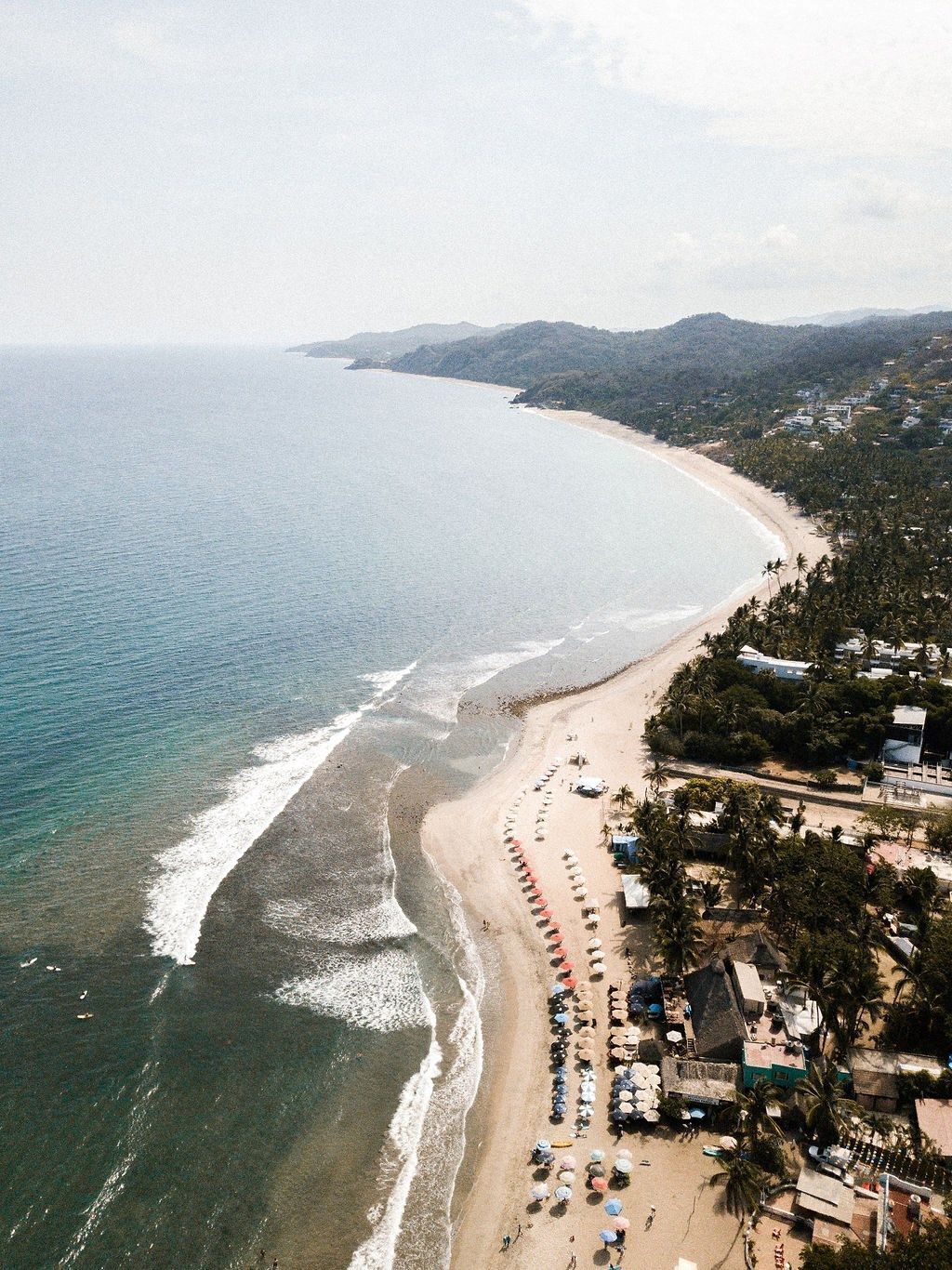An aerial view of a beach with rows of umbrellas and beach chairs, a sandy shoreline, and a backdrop of hills and a clear sky.