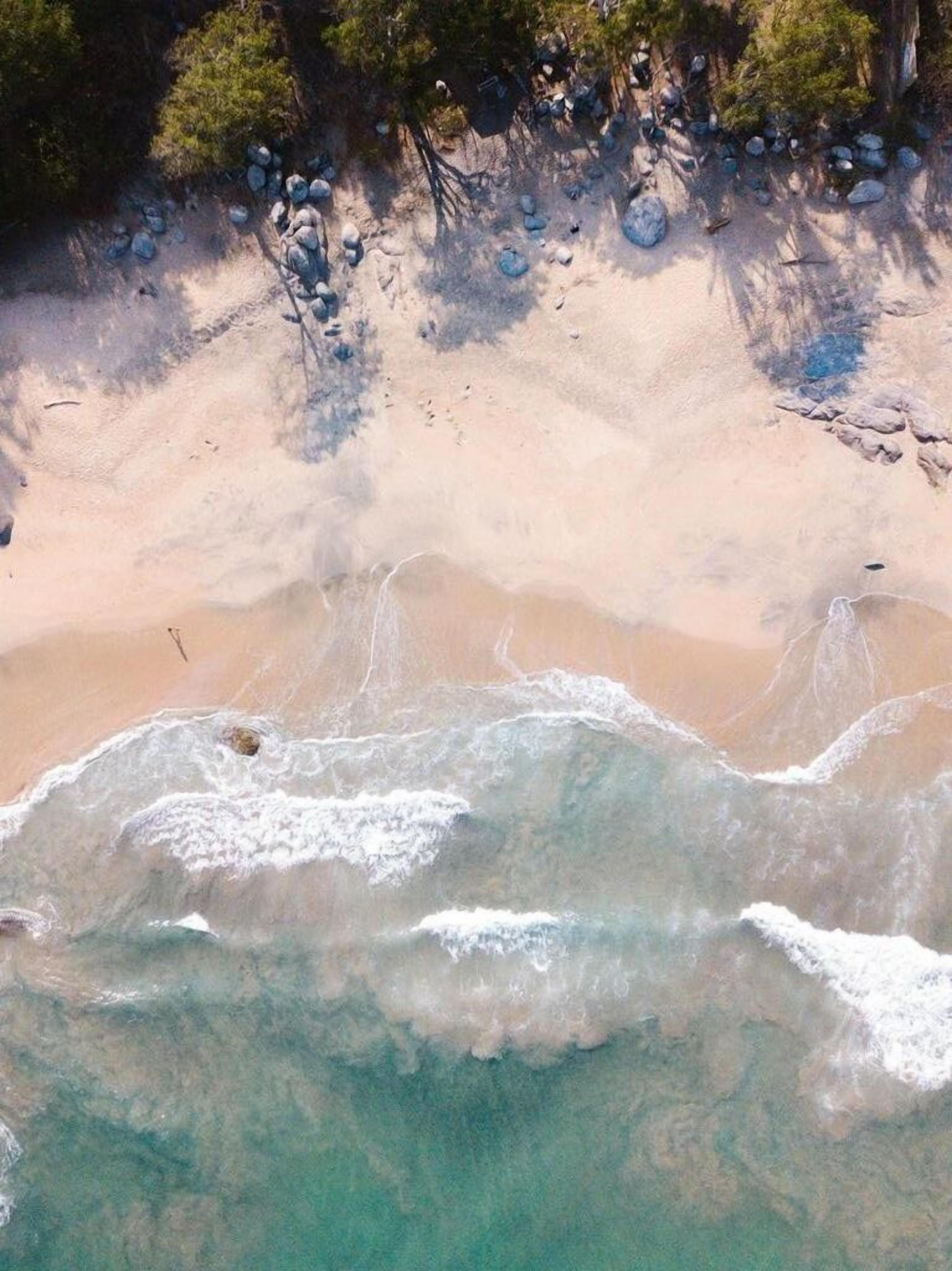 An aerial view of a beach with clear turquoise waters, sandy shore, and scattered rocks.