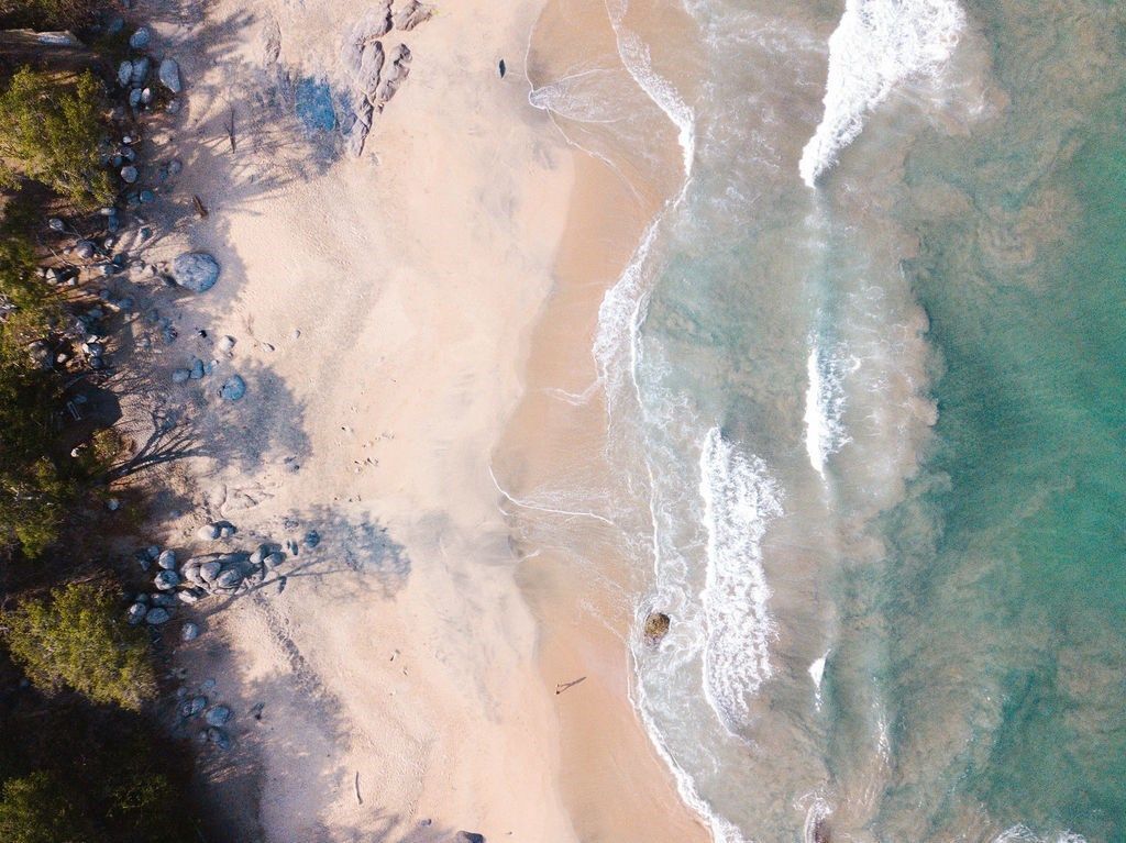 An aerial view of a beach with clear turquoise water, sandy shore, and scattered rocks.