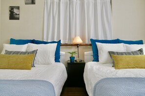 A hotel room with two beds, white bedding, blue and yellow pillows, a bedside table with a lamp, and a small plant.