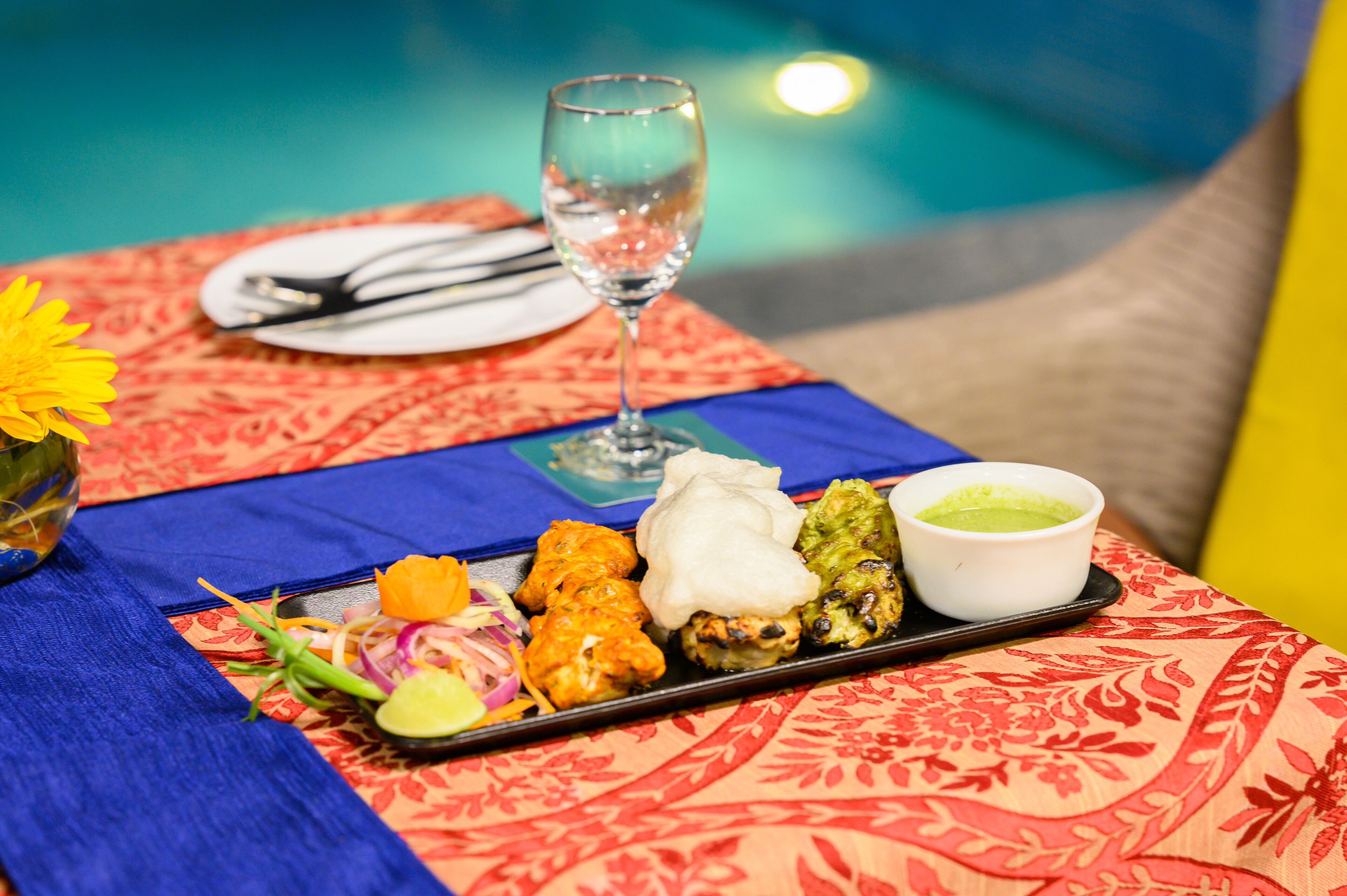 A table with a floral tablecloth, a glass of water, and a tray of food including vegetables, a white sauce, and a green sauce.