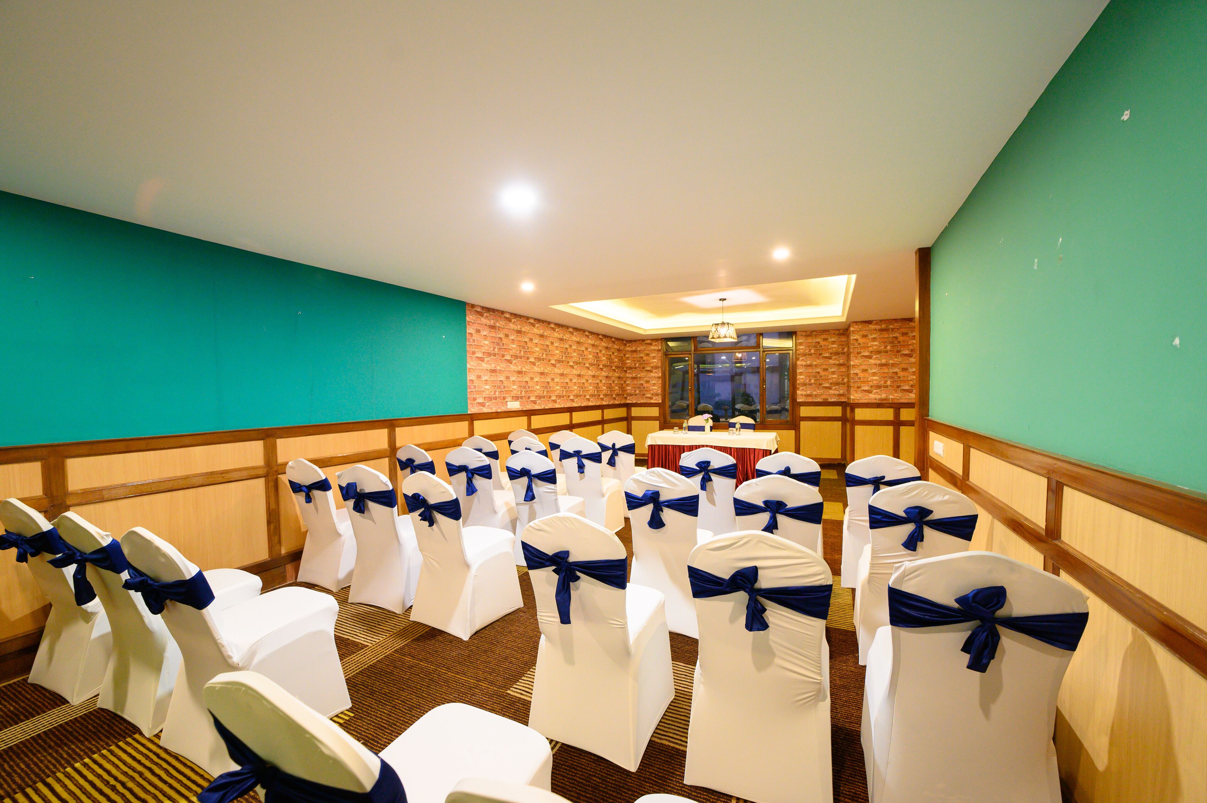 Un salón de conferencias con filas de sillas blancas con cintas azules, una mesa con mantel rojo y una pared de ladrillo.