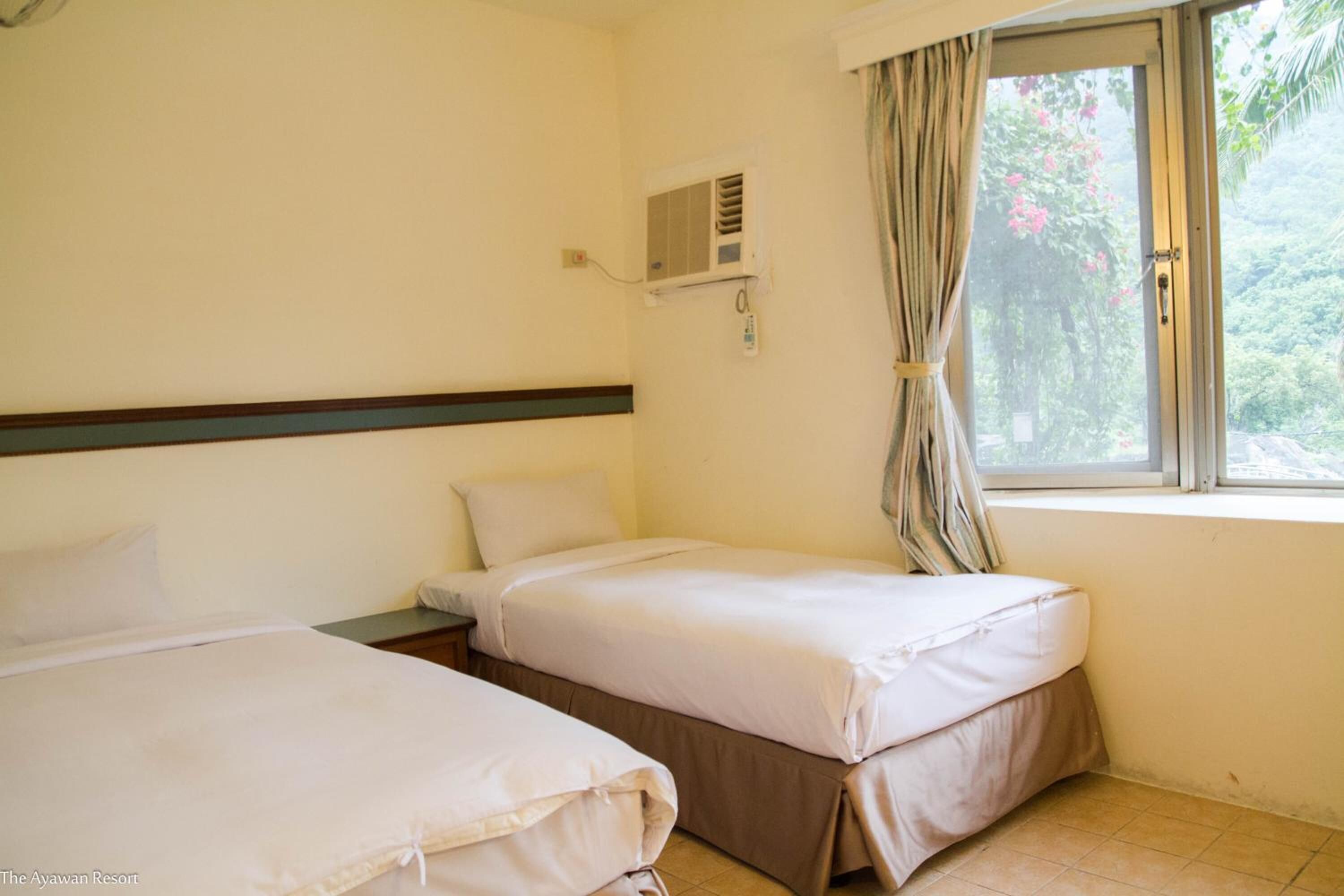 Quarto com duas camas, janela com cortinas, ar condicionado e vista para áreas verdes.