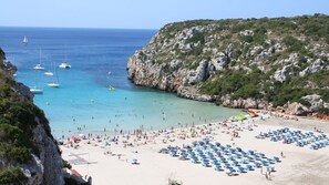 Une plage avec de nombreux parasols et beaucoup de monde, des falaises rocheuses et des bateaux sur l’eau.