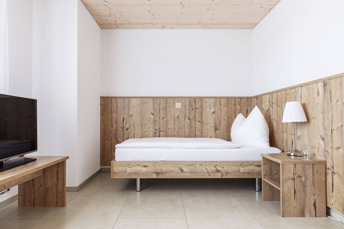 Quarto moderno com cabeceira de madeira, uma cama de casal, mesa de cabeceira e televisão sobre suporte de madeira.