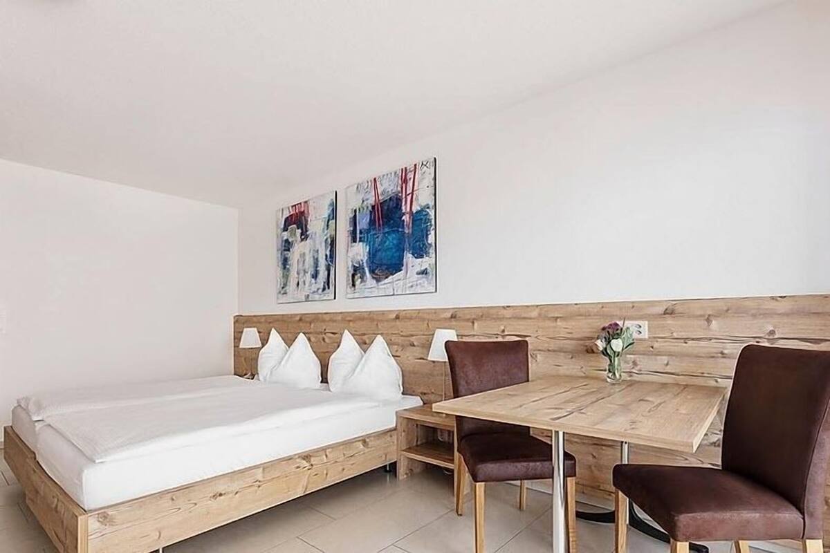 Quarto com cama de madeira, uma mesa de madeira e duas cadeiras. Há duas pinturas penduradas na parede acima da cama.