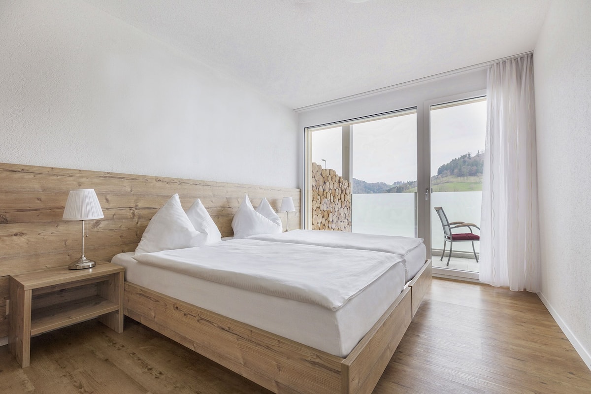 Quarto com cama de madeira, mesa de cabeceira, cadeira junto à janela e vista para o exterior.