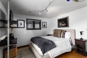 Un dormitorio ordenado con cama, mesitas de noche, un estante en la pared y una ventana con persianas.