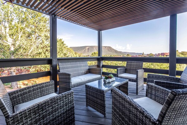 Un espacio de estar al aire libre cubierto con mobiliario de mimbre y una mesa de vidrio, con vistas a un paisaje montañoso.