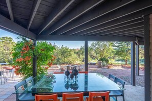 Un área de comedor al aire libre techada, con una mesa y sillas de vidrio, rodeada de follaje y árboles.
